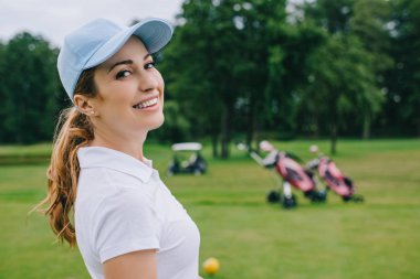 side view of smiling woman in polo and cap looking at camera at golf course clipart