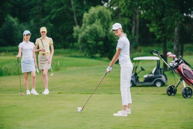selective focus of woman in cap playing golf while friends standing near by at golf course clipart