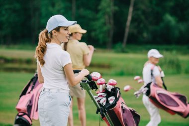 selective focus of women in caps with golf gear walking on green lawn at golf course clipart