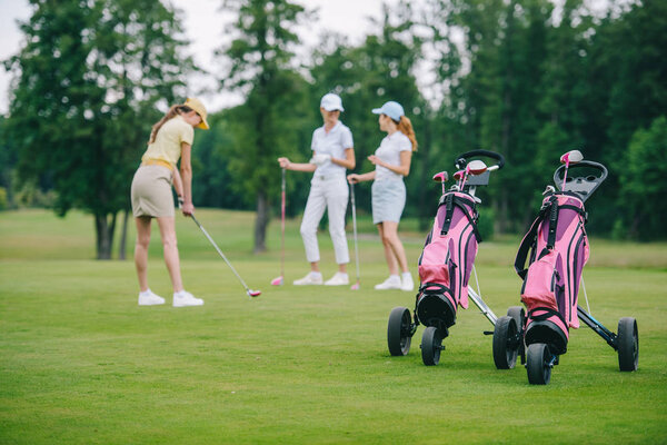 selective focus of golf gear, woman in cap with golf club and friends standing near by at golf course