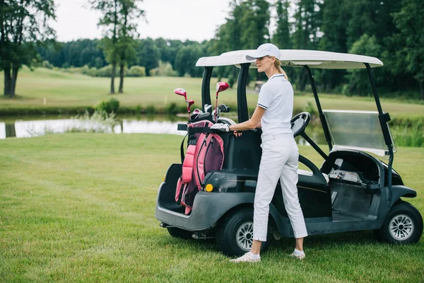 woman in polo and cap with golf gear standing at golf cart at golf course on summer day