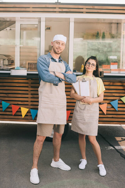 young small business owners in aprons standing with crossed arms and smiling at camera near food truck
