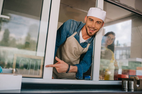 cheerful young man smiling at camera and pointing with finger at sign open in food truck