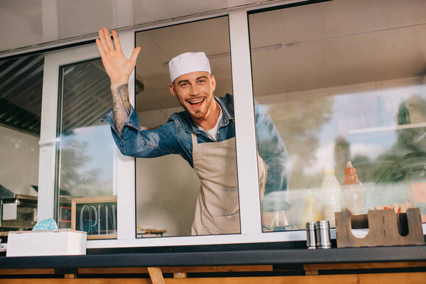 cheerful young man waving hand and smiling at camera while working in food truck