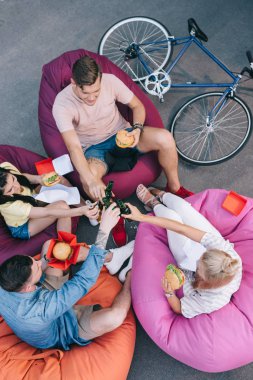 high angle view of friends clinking with bottles of beer and holding fast food on bean bag chairs clipart