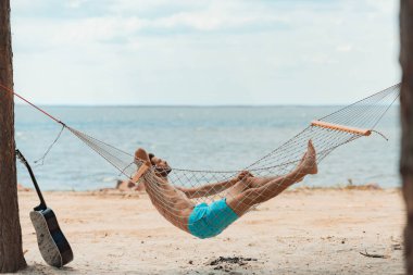 handsome bearded man lying in hammock on beach near the sea clipart