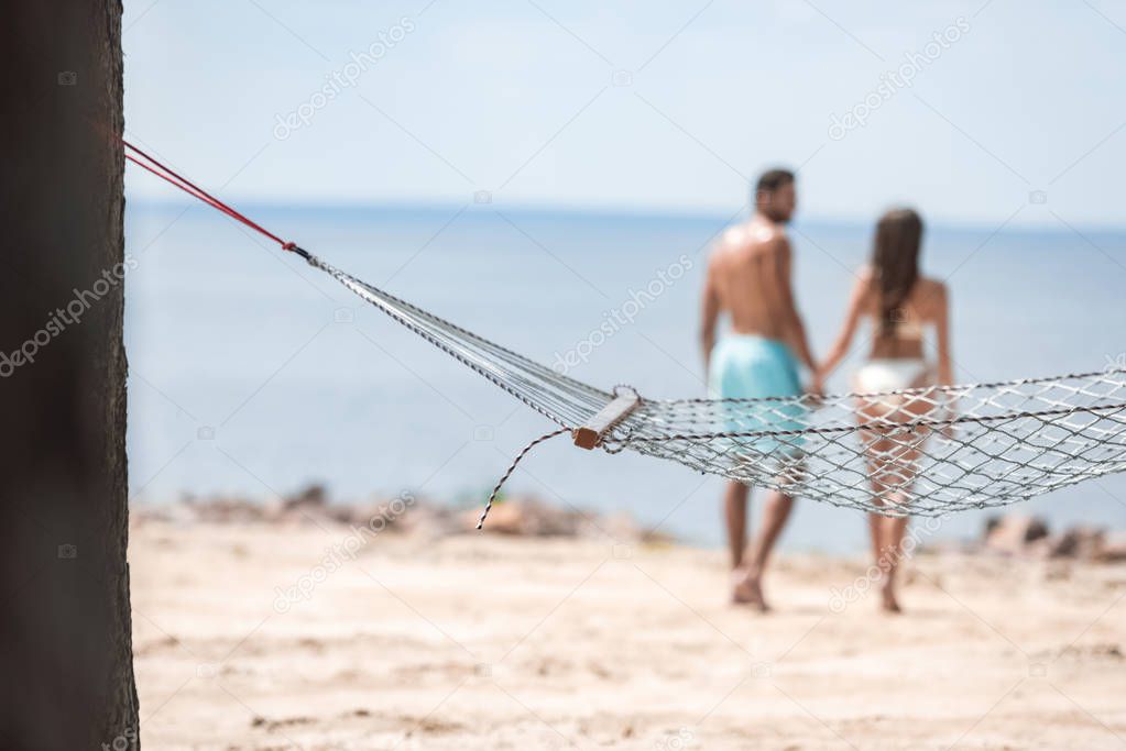 selective focus of couple holding hands and walking on beach with hammock on foreground