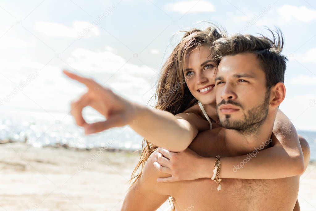 man piggybacking his girlfriend, while she pointing on beach