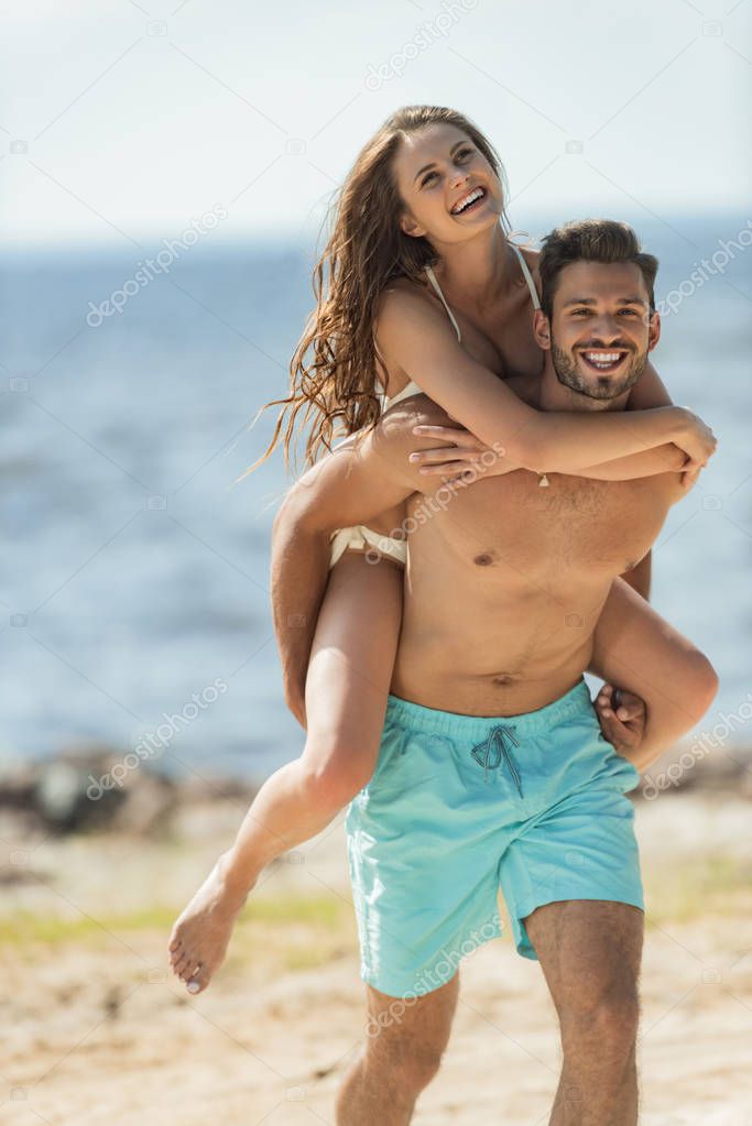 cheerful man piggybacking his smiling girlfriend on seashore