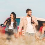 Joven feliz pareja bebiendo cerveza en flor campo mientras su amigos sentado en coche tronco