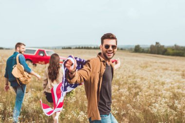handsome young man with united states flag in flower field with friend during car trip clipart