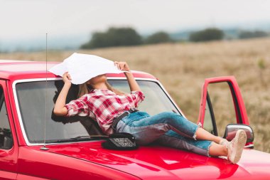 young woman lying on hood of vintage red truck in field and covering face with map in field clipart