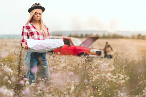 young woman looking at map in flower field while her friends standing near broken car blurred on background