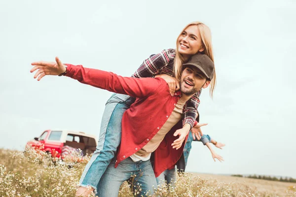 Smiling Young Woman Piggybacking Boyfriend Flower Field Blurred Car Background — Stock Photo, Image