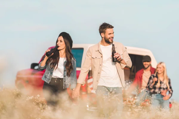 車のトランクに座って彼らの友人間の花畑でビールを飲んで幸せなカップル  — 無料ストックフォト