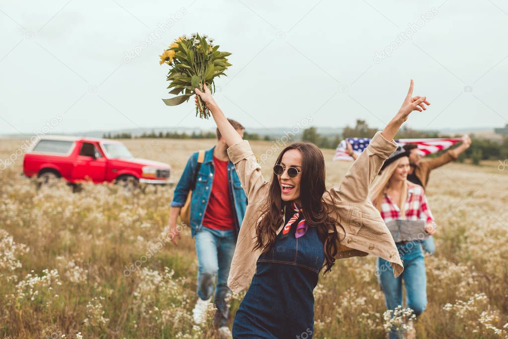 happy young woman holding bouquet and walking by field with friends during trip