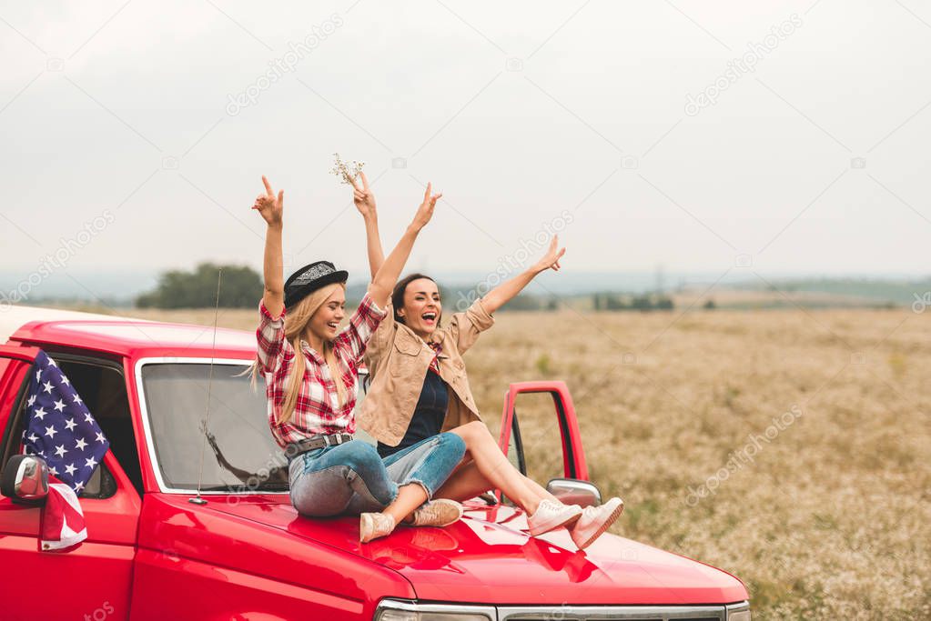beautiful young girlfriends sitting on car hood with raised hands and showing peace signs