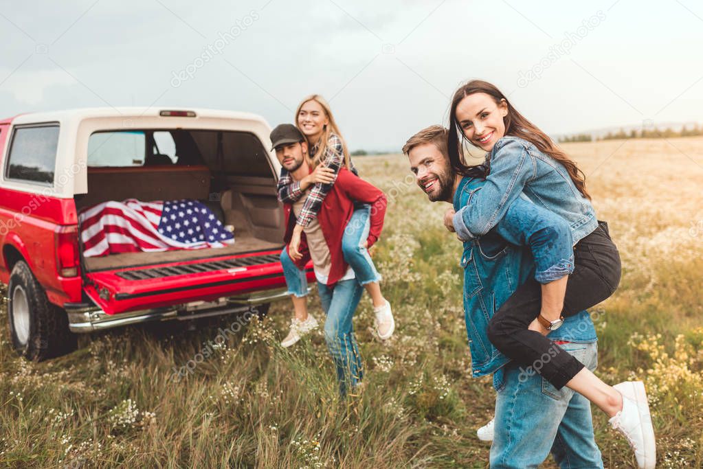 young happy women piggybacking on boyfriends in flower field during car trip