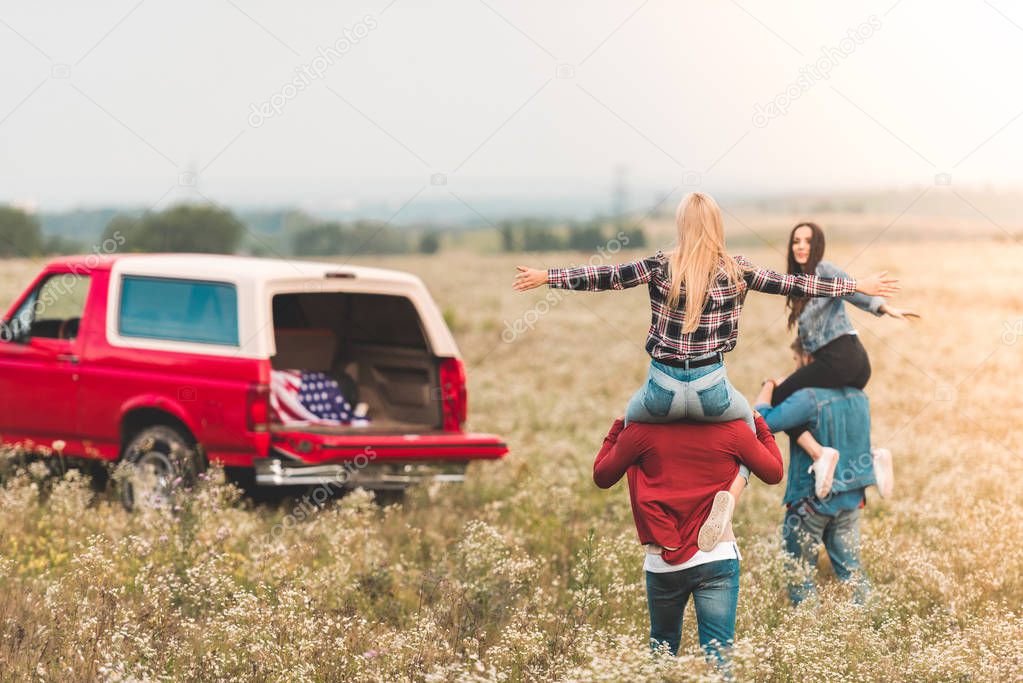young women riding on boyfriends shoulders in field during car trip