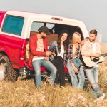Gruppe fröhlicher junger Leute trinkt Bier und spielt Gitarre, während sie es sich im Kofferraum eines Autos auf einem Blumenfeld gemütlich macht