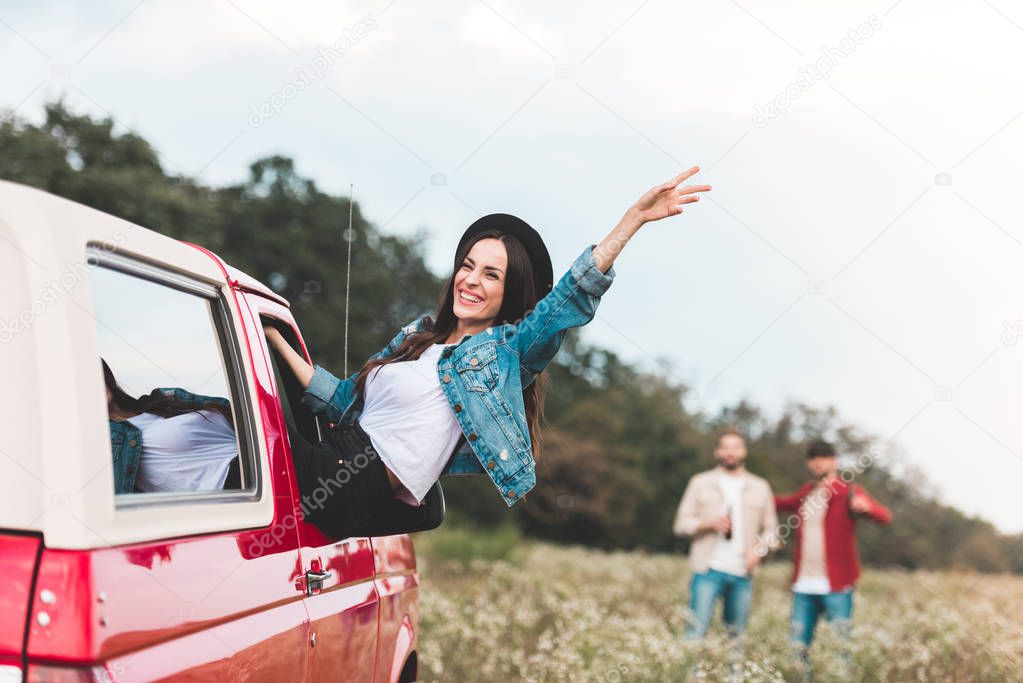 young woman outstretching from car window and raising hand while men standing blurred on background in flower field