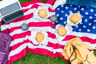 partial view of man lying near american flag with laptop, burgers and coffee to go on green grass clipart