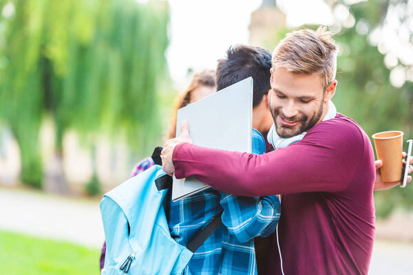 partial view of multicultural students with digital devices hugging in park