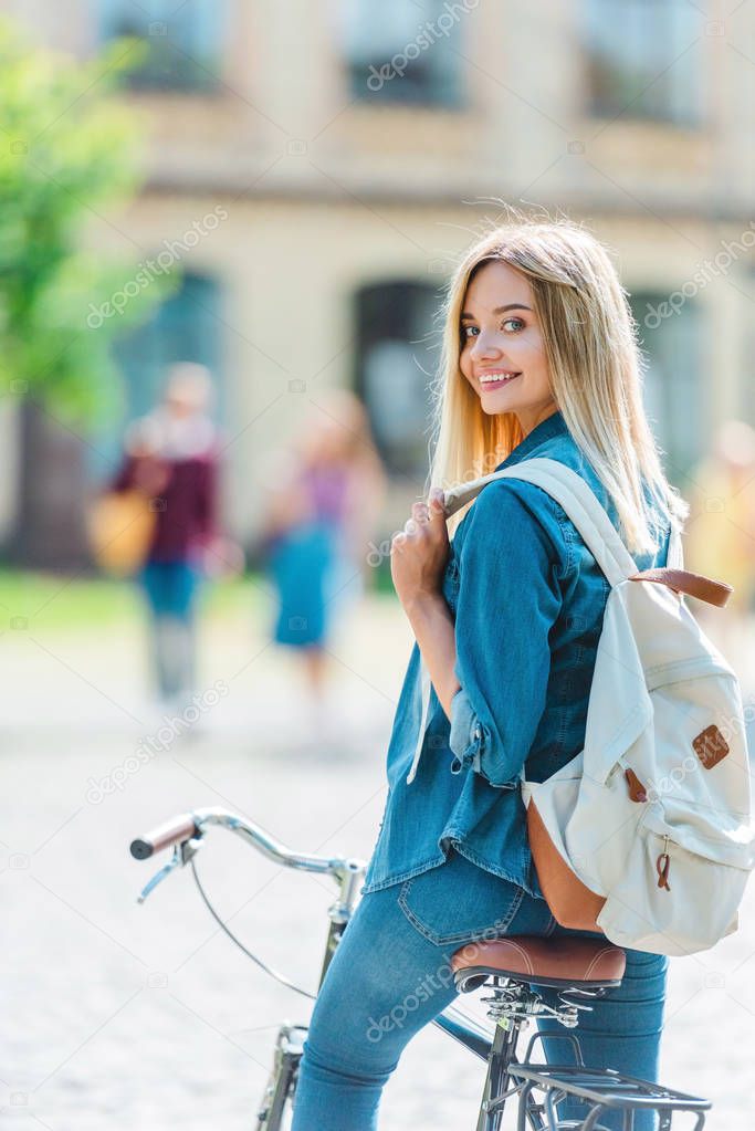 selective focus smiling young student with backpack on bicycle looking at camera on street