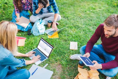 dizüstü bilgisayarlar ve dijital cihazlar Park yeşil çimenlerin üzerinde oturan çok ırklı öğrencilere kısmi görünümü