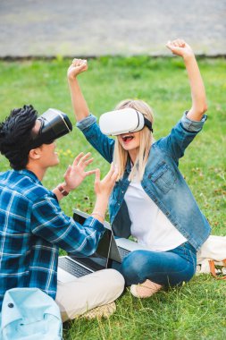 sanal gerçeklik kulaklıklar Park yeşil çimenlerin üzerinde oturan dizüstü bilgisayarlar ile çok kültürlü öğrenci yan görünüm