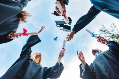 bottom view of happy multicultural graduates with diplomas throwing caps up with blue sky on background clipart