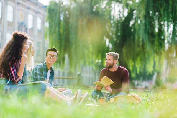 smiling multiracial students resting in park with university on background