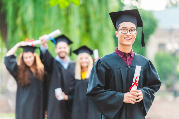 селективный фокус улыбающегося азиатского выпускника с одноклассниками в летнем парке
