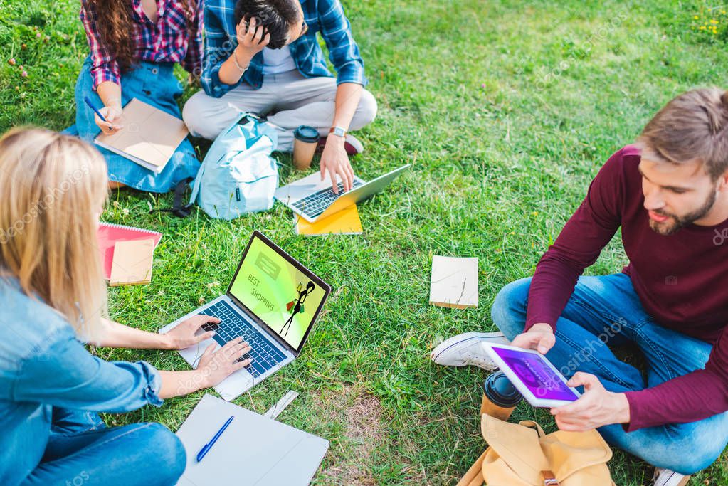 Partial view of multiethnic students with notebooks and digital devices sitting on green grass in park