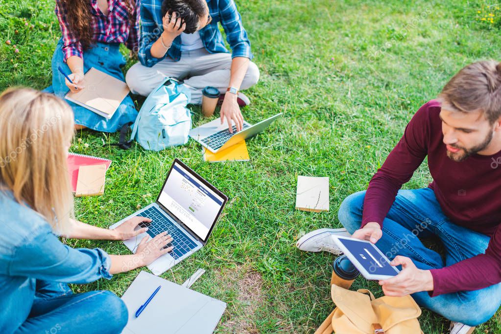 Partial view of multiethnic students with notebooks and digital devices sitting on green grass in park