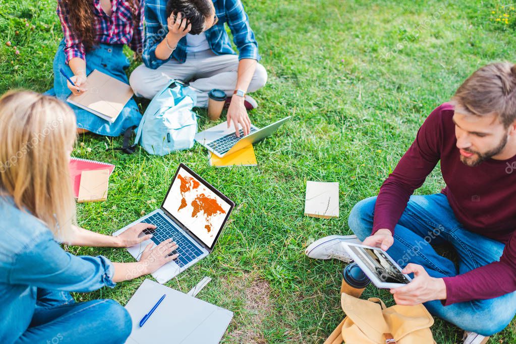Partial view of multiethnic students with notebooks and digital devices sitting on green grass in park
