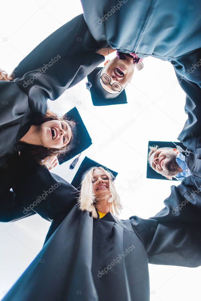 bottom view of cheerful multiracial graduates standing in row and looking at camera