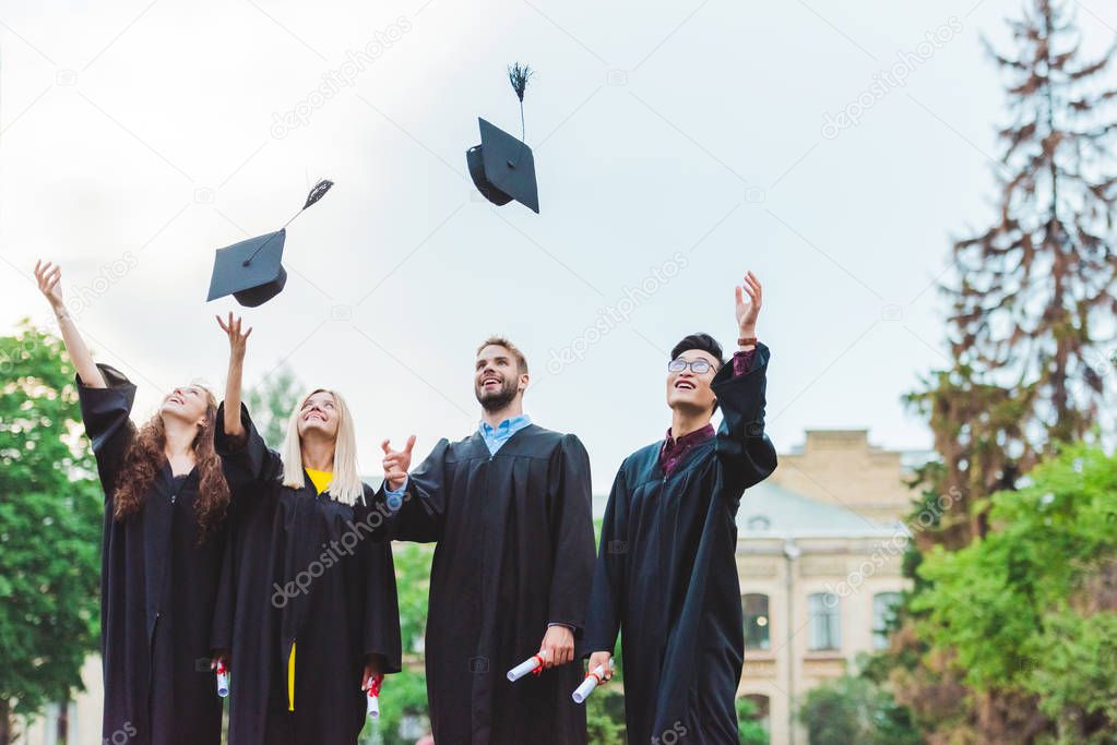 portrait of happy multicultural graduates with diplomas throwing caps up in park