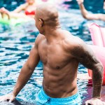 Giovane uomo tatuato vicino a bordo piscina a parlare con gli amici in piscina