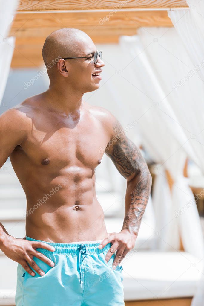 shirtless tattooed muscular man standing near sun loungers and looking away