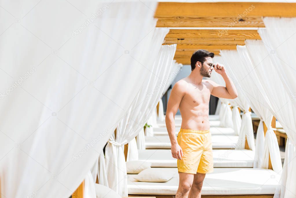 handsome shirtless man in sunglasses standing near sun loungers and looking away