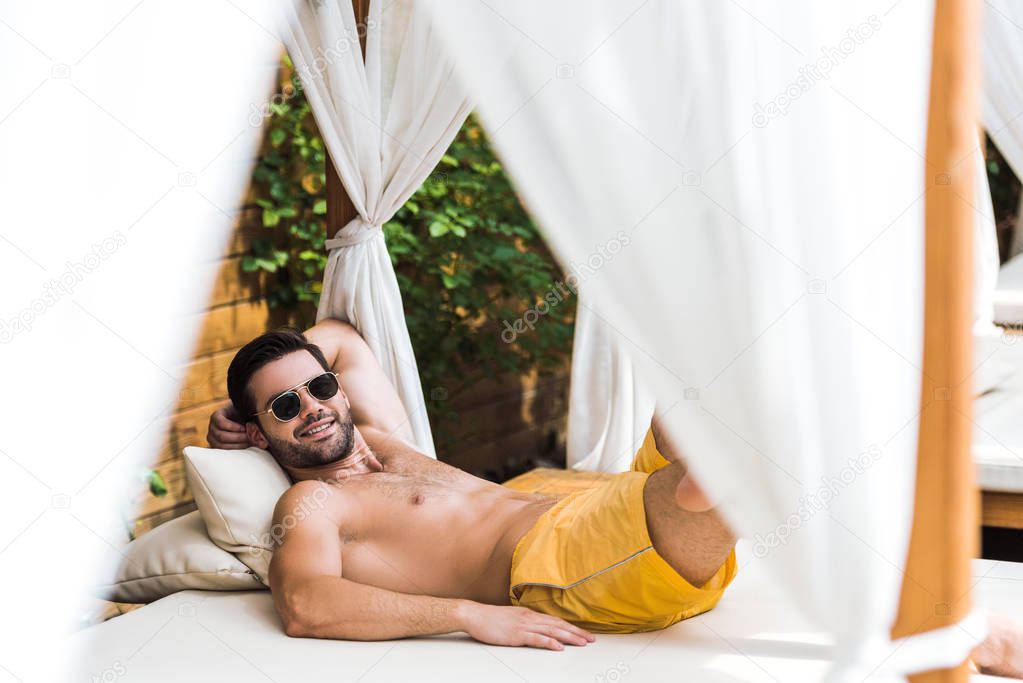 smiling handsome shirtless man in sunglasses lying on sun lounger and looking at camera
