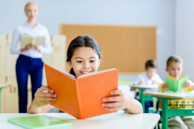 beautiful smiling schoolgirl reading book at classroom during lesson