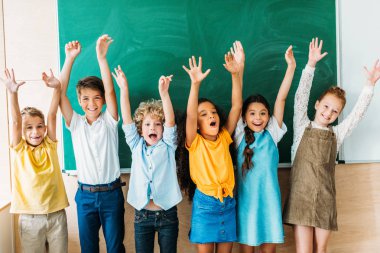 adorable happy schoolchildren with raised hands standing in front of blank chalkboard clipart