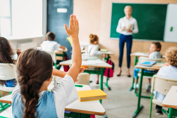 rear view of schoolgirl raising hand to answer teachers question during lesson