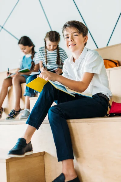 Gelukkig Schooljongen Schrijven Notitieboekje Camera Terwijl Zijn Klasgenoten Zitten Achter — Stockfoto