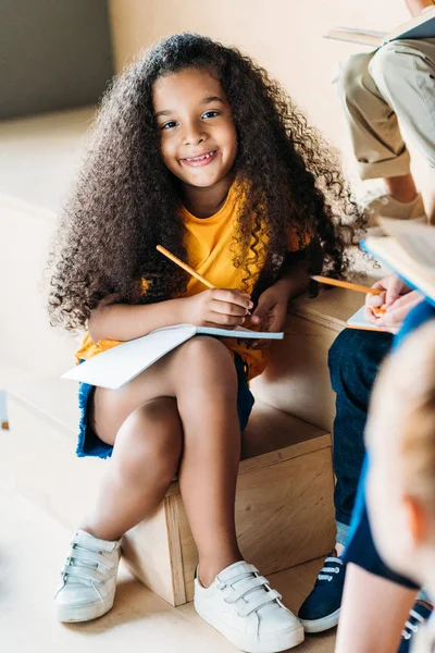 Schattige Lachende Afro Amerikaanse Schoolmeisje Schrijven Notitieboekje Camera Kijken — Stockfoto