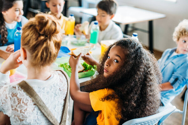 african american schoolgirl taking lunch at school cafeteria with her classmates and looking at camera