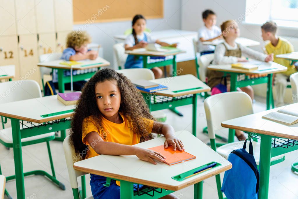 beautiful bored african american schoolgirl sitting on lesson with classmates blurred on background