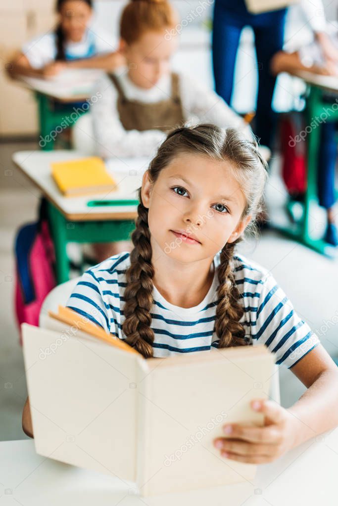 beautiful little schoolgirl with book looking at camera during lesson at school